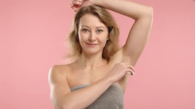 Kişisel bakım anında, genç beyaz bir kadın kollarının altına deodorant sürüyor, pembe bir arka plana havluyla sarılıp günlük rutininde hijyen ve tazeliği vurguluyor.. 
