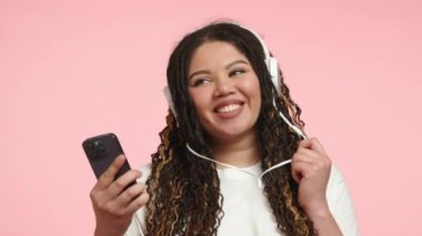 Kıvırcık saçlı neşeli genç bir bayan dijital müzik akışı deneyimine dalmış, kablosuz kulaklığı ve akıllı telefonu kullanarak, teknolojiye ve modern yaşam tarzına olan aşkını gösteriyor.