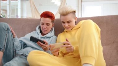 Eşsiz saç stilleri olan iki arkadaş ve günlük giysiler kanepede gezici oyunlar oynuyorlar, içeride gülüyorlar ve etkileşime geçiyorlar. Eğlenceli ve rahat bir zamanda teknoloji ve arkadaşlıktan hoşlanırlar.