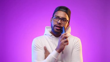 Telleri olan düşünceli Afro-Amerikalı erkek telefonuna büyüleyici bir mor arka plan karşısında heyecanlı bir şekilde bir mesaj yazmadan önce ne yazacağını düşünüyor. Kamera 8K HAM. 