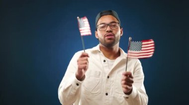 Bir adam Amerikan bayraklarıyla dans ederken mutluluk saçar, kutlama ve vatansever ruhu simgeler derin mavi bir arka plana karşı bir sevinç anında. Kamera 8K HAM. 