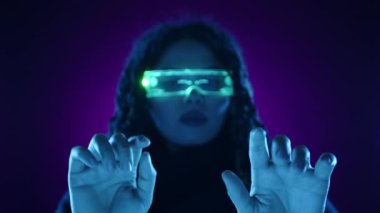 Neon ışıklı gözlüklü bir kadının canlı mor ve deniz mavisi aydınlatma altında holografik bir arayüzle etkileşime geçtiği ileri teknoloji ve yenilik içeren fütürist bir sahne.