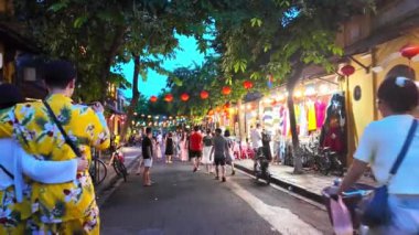 HOI AN, VIET NAM - 17 Haziran 2024: Kalabalık turistler Hoian antik kasabasında akşamları hayattan zevk alıyorlar, mavi gökyüzünde fenerleri olan eski evlerden rengarenk, yürüyen veya güzel sokak mirasında bisiklet süren insanlar