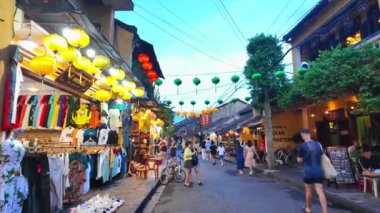 HOI AN, VIET NAM - 17 Haziran 2024: Kalabalık turistler Hoian antik kasabasında akşamları hayattan zevk alıyorlar, mavi gökyüzünde fenerleri olan eski evlerden rengarenk, yürüyen veya güzel sokak mirasında bisiklet süren insanlar