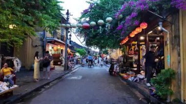HOI AN, VIET NAM - 17 Haziran 2024: Kalabalık turistler Hoian antik kasabasında akşamları hayattan zevk alıyorlar, mavi gökyüzünde fenerleri olan eski evlerden rengarenk, yürüyen veya güzel sokak mirasında bisiklet süren insanlar