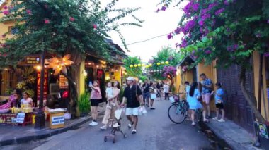 HOI AN, VIET NAM - 17 Haziran 2024: Kalabalık turistler Hoian antik kasabasında akşamları hayattan zevk alıyorlar, mavi gökyüzünde fenerleri olan eski evlerden rengarenk, yürüyen veya güzel sokak mirasında bisiklet süren insanlar