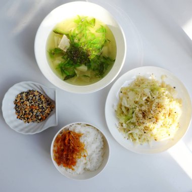 Buharda pilav, tofu ve sebze çorbası karıştırılmış yeşil fasulye, bambu filizleri ve beyaz bir masanın üzerindeki kırmızı biber sosu, bir pencereden gelen ışıkla aydınlatılmış.