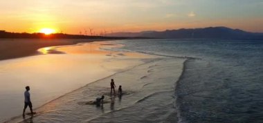 Quy Nhon sahilinde gün batımı, Binh Dinh, Vietnam. Bir baba ve üç çocuğunun plajda yürüyüşleri, yürek ısıtan sahneler. Rüzgar türbinleri, dingin deniz burunlarında yenilenebilir enerji.