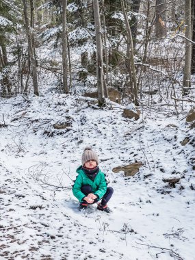 Ormanda yürürken çıplak ağaçlara karşı karada çömelirken karla oynayan kalın giysiler giyen bir çocuk.