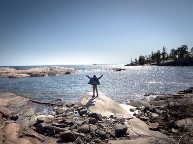 Güneşli bir günde kayalık sahilde dikilirken kollarını kaldırıp deniz manzarasına bakan kaygısız çocuk.