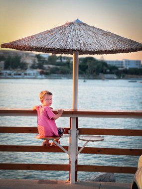 Gün batımında deniz manzarasının önündeki sazdan çatı altında oturan sevimli çocuk portresi