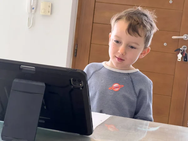可爱的白人男孩在家里上网学习的时候 一边认真地看着桌上的数码平板电脑 — 图库照片