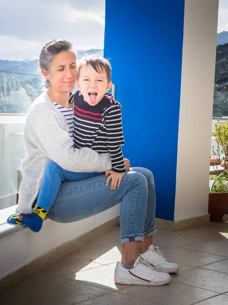 バルコニーで笑顔の母の膝に座っている間に舌を突き出す遊び心のあるかわいい白人少年の肖像画 — ストック写真