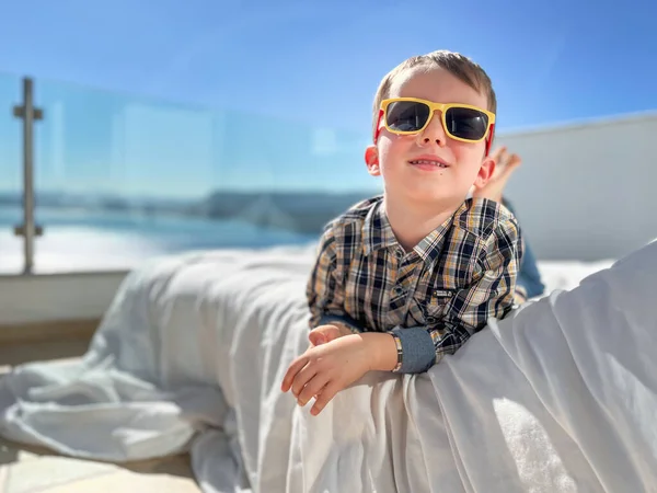 Portret Uroczego Chłopca Noszącego Okulary Przeciwsłoneczne Odpoczywającego Łóżku Przed Jasnym — Zdjęcie stockowe