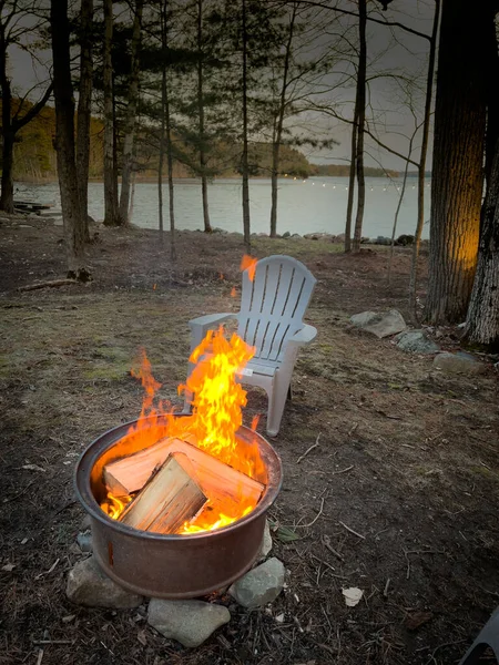 日落时用篝火在森林里的树木和湖面上燃烧的原木看空椅 — 图库照片