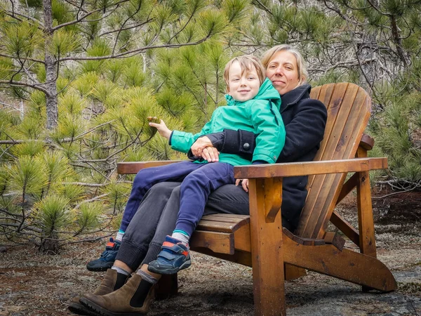休暇中に森の中で成長する緑豊かな植物に対して木製の椅子に座って冬のコートを着て笑顔の母親と息子 ストック写真
