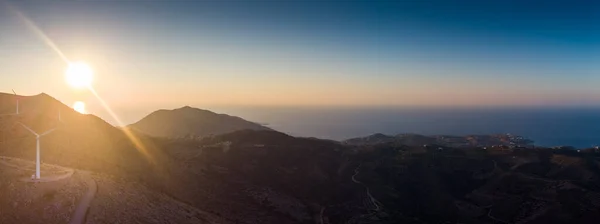 山の上の風力タービンのパノラマビューと日没時の空の太陽に対する風景の景色 ストックフォト