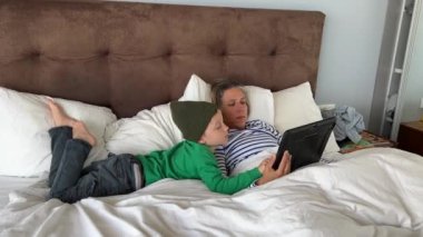 Anne ve oğlu evde vakit geçiriyorlar. Genç bir erkek ve kadın yatakta dijital tablet kullanıyorlar.