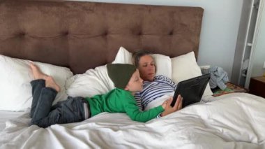 Anne ve oğlu evde vakit geçiriyorlar. Genç bir erkek ve kadın yatakta dijital tablet kullanıyorlar.