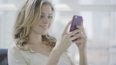 Genç sarışın kadın evde cep telefonuyla mesajlaşıyor.