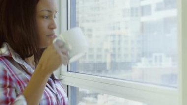 Kareli gömlek giyen güzel siyah kadın panoramik pencerede duran beyaz bardaktan içecek içiyor.