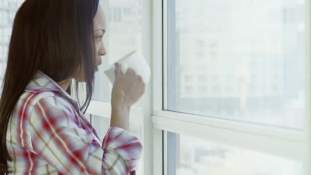 漂亮的黑人女人穿着格子衬衫 站在全景窗前喝着白杯里的饮料 — 图库视频影像
