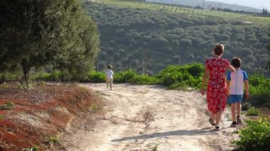 Anne ve iki oğlu kırsalda yolda yürüyor.