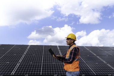 Güvenlik kaskındaki erkek mühendisin portresi güneş istasyonundaki bir güneş paneli çalışmasını bilgisayarla kontrol ediyor.