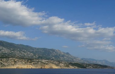 Mavi gökyüzünde yüzen beyaz bulutlar, deniz kıyısındaki kayalıkların tepesinde.