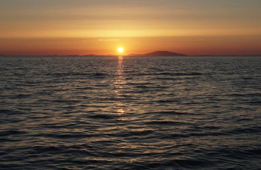 Deniz yüzeyinin soluk ve pastel renkleri. Ufukta küçük dalgalar ve gün batımı var.