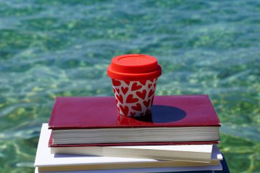 Yığılmış kitapların üzerinde turkuaz deniz altyapısı olan kalp şeklinde bir kahve fincanı.