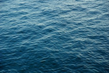 Deniz suyunun tepesinde küçük dalgalar ve ışık yansıması olan parlak eğimli mavi deniz yüzeyi.