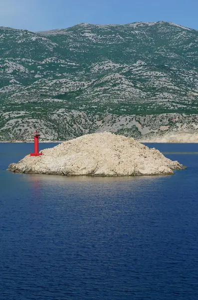 Anakaranın önünde küçük kırmızı bir deniz feneri ve arka planında dağ olan küçük kayalık bir ada.