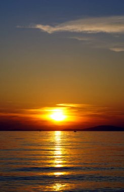 Hırvatistan 'ın Pag adasında mavi gökyüzü ve parlak dalgalı deniz yüzeyi ile canlı turuncu gün batımı