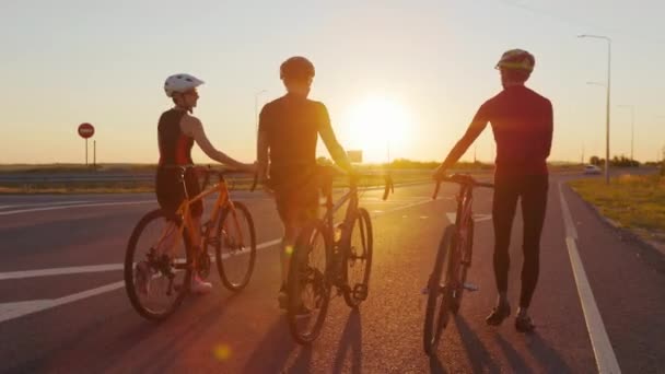 两名男子和一名妇女穿着运动服 戴着头盔 一边聊天 一边微笑 一边骑自行车出去散步 青少年在户外运动中享受夏日日落 — 图库视频影像