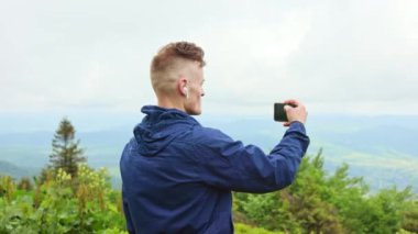 Kulaklıklı yürüyüş yapan, tepelerin fotoğrafını çeken genç, sportif bir erkek. Erkek, spor giyer, tepede durur, doğanın güzelliğine hayran olur. Seyahat ve yürüyüş kavramı.