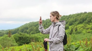 Rüzgarlı havada yürüyüş yapan çekici genç bir kadının yan görüntüsü. Sarışın, kadın turist elinde telefonla tepelerin fotoğrafını çekiyor, manzaraya hayran. Turizm ve yürüyüş kavramı.