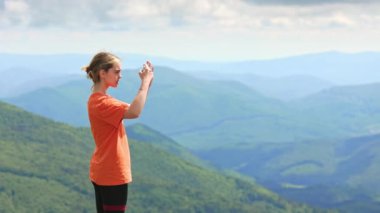 Kadın turistin ayakta durup, dağların videosunu çekerken yan görüntüsü. İnce, sarışın kadın ayakta, elinde telefonla, dağların güzelliğine hayran. Seyahat ve yürüyüş kavramı.