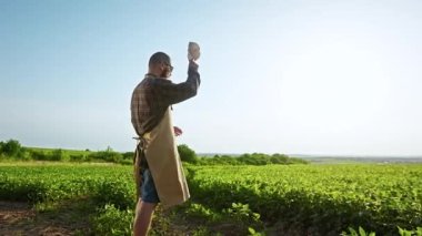 Kırsal kesimde tarlada çalışan genç bir erkek çiftçinin arka görüntüsü. Sahada duran sakallı bir adam, sıcak ve güneşli havada şapkasını takıyor. Bitkilerle ilgilenme kavramı.