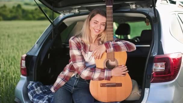 美しい女性の側のビューの車のトランクに座って 抱擁ギター ブロンドの女性笑顔 カメラを見て 車で移動し お楽しみください 旅の概念 — ストック動画