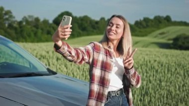 Sarışın, ekose gömlek ve kot pantolon giyen, selfie çeken genç bir bayan. Arabayla seyahat eden neşeli bir bayan, gülümsüyor, akıllı telefon tutuyor, kullanıyor. Modern yaşam tarzı kavramı.