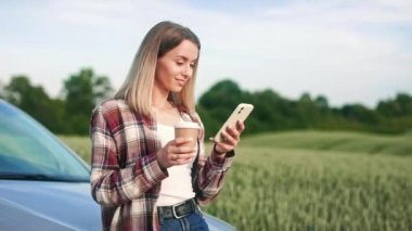 Arabasında oturan genç, sarışın bir kadın, akıllı telefon kullanıyor. Ekose gömlek ve kot pantolon giyen neşeli bayan, mesajlaşıyor, sohbet ediyor, kahve içiyor. Modern yaşam tarzı kavramı.