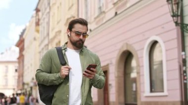 Sırt çantası taşıyan meşgul turistin yan görüntüsü, caddede yürüyen gong sesi. Sakallı, güneş gözlüğü takan, akıllı telefon kullanan yakışıklı bir erkek. Modern yaşam tarzı kavramı.