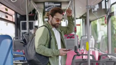 Sırt çantası ve güneş gözlüğü takan, otobüsle giden, ayakta duran meşgul bir adamın yan görüntüsü. Sakallı, kulaklık takan, akıllı telefon tutan yakışıklı bir erkek. Toplu taşıma kavramı.