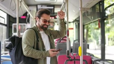 Side view of handsome man going by bus, standing, holding, using smartphone. Brunette male with earphones wearing sunglasses, texting, scrolling. Concept of public transport.