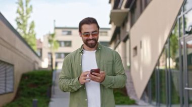 İş adamının yürümesini, sokağa çıkmasını, elinde tutmasını, akıllı telefon kullanmasını. Sakallı, güneş gözlüklü, modaya uygun kıyafetler giyen, kayan, gülümseyen bir erkek. Modern yaşam kavramı.