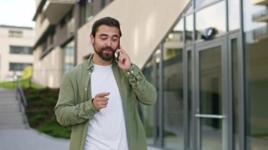 Sakallı bir adam sokakta yürüyor, akıllı telefon kullanıyor, konuşuyor. Yakışıklı, esmer erkek, beyaz tişört ve tişört giyiyor. Modern şehir yaşam tarzı kavramı.