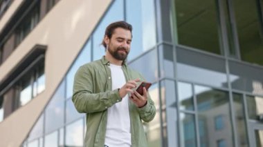 Yakışıklı, esmer adamın sokakta akıllı telefon kullanışının yan görüntüsü. Beyaz tişört ve tişört giyen erkek, cam binanın yanındaki sokakta duruyor. Modern, kentsel yaşam tarzı kavramı.