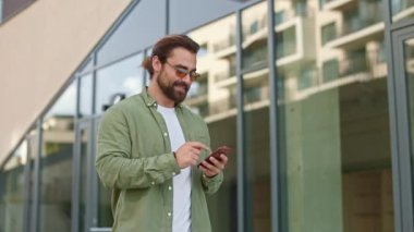 Güneş gözlüğü takan, sokakta yürüyen, elinde akıllı telefon tutan meşgul bir adamın yan görüntüsü. Sakal okuyan, kaydıran, mesaj atan, gülümseyen bir iş adamı. İş ve modern hayat kavramı.
