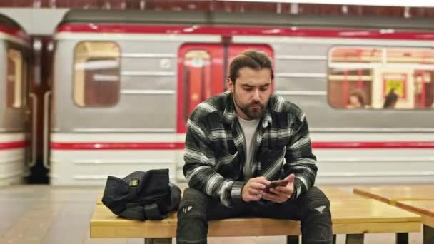 在地铁里等着的一个大忙人坐在长椅上 留着胡子的布鲁内特男性 使用智能手机 发短信 现代城市生活的概念 — 图库视频影像